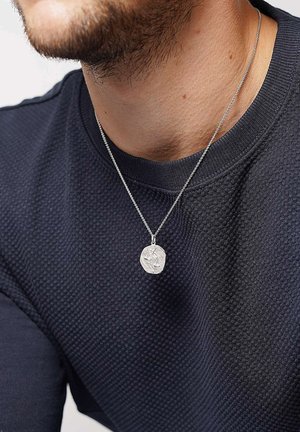 Silberne Halskette mit einem texturierten runden Anhänger, der geprägte Schuppen zeigt, aufgehängt an einer feinen Kette und über einem nav blauen texturierten Pullover getragen.