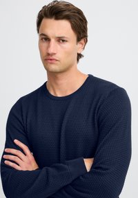Navy blue textured sweater with a round neckline and long sleeves. The fabric features a subtle pattern and ribbed cuffs. Model is posing with arms crossed.