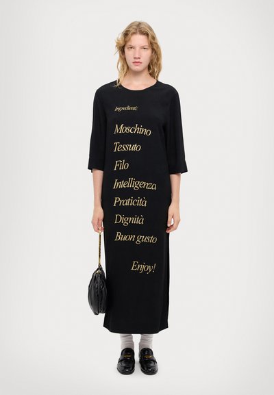 Long black dress with gold text, three-quarter sleeves, loose fit, below knee length. Worn with a small black bag and dark shoes.