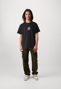 Camiseta de algodón negra con un gráfico colorido de una cereza y la letra D en el frontal, combinada con pantalones cargo verde oliva y zapatillas blancas.