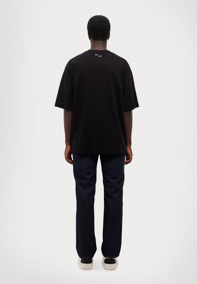 Svart bomullströja med en avslappnad passform, korta ärmar och en subtil detalj på baksidan; kombinerad med mörkblå jeans och svart-vita sneakers.
