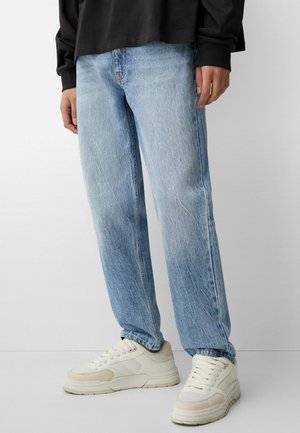 Oseba v svetlo modrih ravnih hlačah, belih športnih čevljih in dolgem črnem zgornjem delu, stoji ob enotnem ozadju.