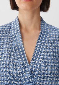 Blå mönstrad blus med V-ringning. Tyget har en geometrisk motiv i nyanser av blått och vitt, med en slät textur.