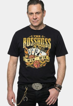 Mężczyzna noszący czarną koszulkę „The Rosshoss Win Win” z wężem, czterema asami, kośćmi do gry, paskiem z klamrą w kształcie byka, bransoletkami i łańcuchem z podkową.