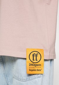 Šviesiai rožinė marškinėliai, pagaminti iš minkštos medvilnės, su tiesia apatine dalimi. Juos puošia ryškiai oranžinė etikete su juodu logotipu ir tekstu ant šono.