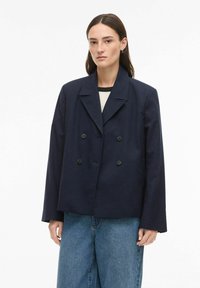 Femme aux cheveux bruns lisses portant un blazer bleu marine foncé oversize à double boutonnage et un jean bleu ample sur fond blanc.