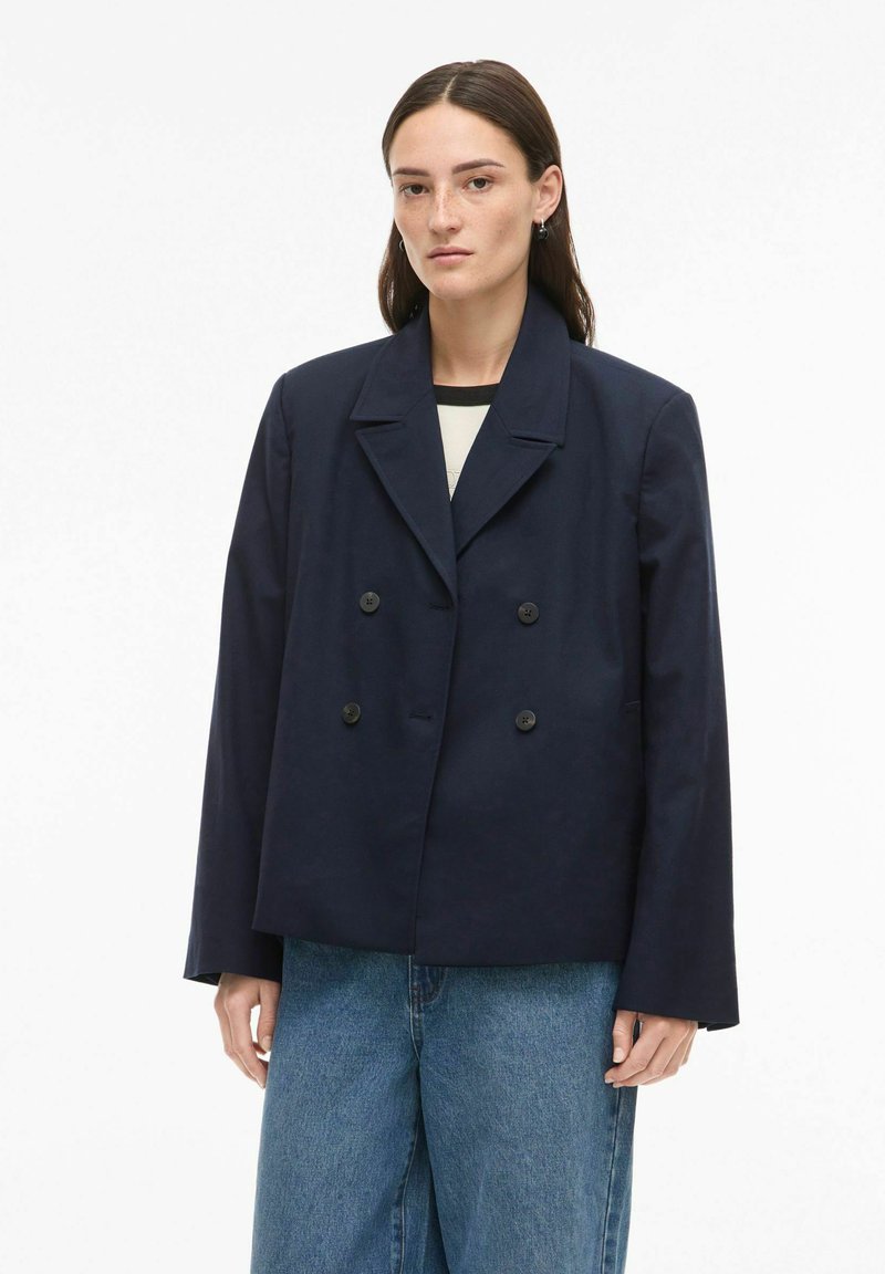 Femme aux cheveux bruns lisses portant un blazer bleu marine foncé oversize à double boutonnage et un jean bleu ample sur fond blanc.