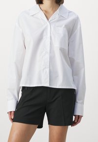 Camicetta bianca a maniche lunghe, con bottoni e taschino sul petto, abbinata a pantaloni corti neri con bottoni laterali e una texture di tessuto liscia.