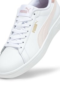 Weiße Sneakers mit einer glatten synthetischen Oberfläche, ausgestattet mit einem rosa PUMA-Seitenstreifen, goldenem Logo und weißen Schnürsenkeln auf einer hellgrauen Außensohle.