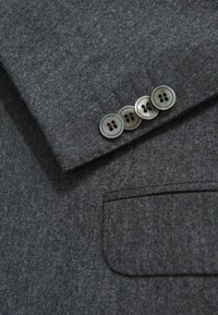 Giacca di lana grigia in primo piano con una superficie texturizzata, caratterizzata da quattro bottoni rotondi scuri e una tasca sul petto.