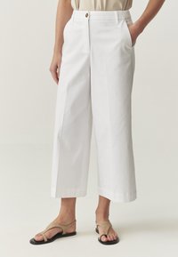 Pantaloni bianchi a vita alta con una vestibilità comoda, dotati di chiusura con bottone frontale, tasche laterali e un tessuto di cotone morbido. Abbinati a sandali beige.