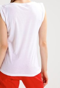 Biała bezrękawnik z okrągłym dekoltem i subtelnym wykończeniem ramion. Gładka bawełniana tkanina, noszona z czerwonymi spodniami.