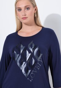 Marineblauwe T-shirt met lange mouwen, voorzien van een glanzend zilveren vlamontwerp met zwarte rhinestones en het woord "LOVELY" in een gebogen lettertype.