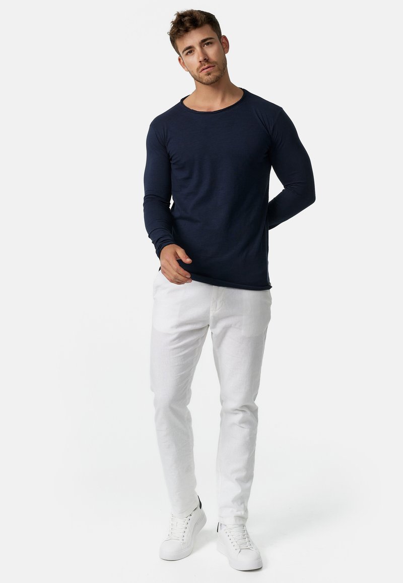 Jeune homme portant une chemise bleu marine à manches longues, un pantalon blanc et des baskets blanches, debout avec une main derrière le dos sur un fond uni.