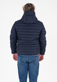 Marineblauwe gewatteerde jas met een capuchon, voorzien van horizontale stiksels en een rits aan de voorkant. G gecombineerd met blauwe denim jeans.