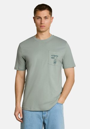 Man wearing light green short-sleeve shirt with chest pocket printed "VITAMIN SEA" and a small bottle illustration, paired with blue jeans.