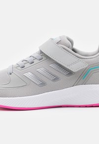 Baskets de sport gris clair avec tissu en mesh, rayures argentées en angle, sangle velcro, semelle blanche et accent rose sur la semelle extérieure.