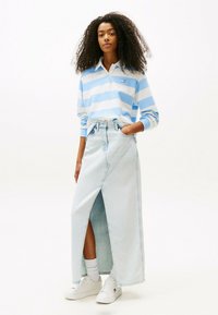 Polo shirt à manches longues rayé bleu et blanc, associé à une jupe maxi en denim clair taille haute avec une fente à l'avant. Baskets blanches aux pieds.