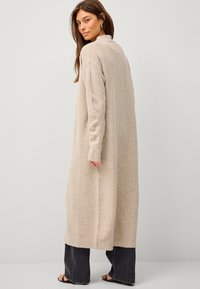 Langer beiger Strickcardigan mit hohem Kragen, gerippten Bündchen und seitlichen Schlitzen. Kombiniert mit dunklen, weit geschnittenen Jeans, die eine strukturierte Oberfläche aufweisen.