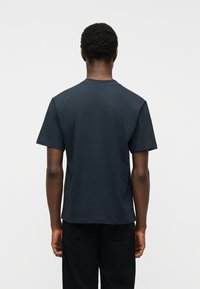 Άνδρας που φοράει μια απλή σκούρα μπλε κοντομάνικη μπλούζα και μαύρο παντελόνι, με την πλάτη προς το μέρος μας, μπροστά σε ένα ανοιχτό γκρι φόντο.