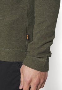 Olivgrön långärmad skjorta med en texturerad tyg, som har synliga sömdetaljer och en liten orange "BOSS"-etikett på sidan.