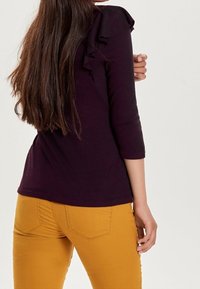 Fialový top s volánovými detaily na ramene, tři čtvrtinové rukávy, spárovaný s hořčicově žlutými kalhotami s zadními kapsami a slim fit.