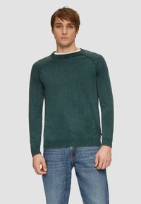 Groene katoenen trui met een ronde hals, lange mouwen en geribde manchetten; heeft een zachte textuur en een ontspannen pasvorm. Gemaakt in combinatie met een blauwe spijkerbroek.