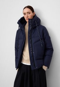 Kobieta w granatowej, pikowanej kurtce z wysokim kołnierzem i kapturem na sznurek, w połączeniu z beżowym swetrem i czarną spódnicą, stojąca przed białą ścianą.