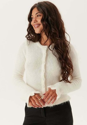 Hvid strikket cardigan lavet af blødt, fluffy materiale. Har en rund halsudskæring, knaplukning med små perler og flæseærmer.