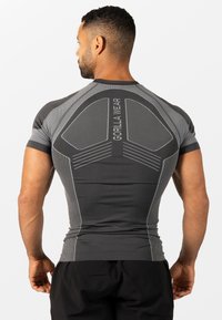 Moški, ki nosi oprijeto sivo-črno športno majico blagovne znamke Gorilla Wear, prikazan od zadaj, stoji pred belo enobarvno ozadje.