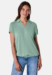 Hellgrünes Polo-Shirt mit kurzen Ärmeln, V-Ausschnitt und aufgerollten Bündchen. Hergestellt aus glattem Stoff mit lockerer Passform.