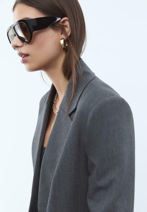 Grå skræddersyet blazer lavet af tekstureret stof, oversized sorte solbriller, guld hoop øreringe og en guld kæderekke.