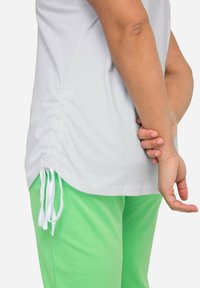 Camiseta blanca de algodón con detalles fruncidos en los laterales y cordones, combinada con pantalones jogger verdes. Diseño sencillo, corte relajado y textura suave.