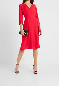Robe rouge à manches en dentelle trois-quarts, avec une taille cintrée et une jupe fluide. Portée avec des sandales noires à talons hauts et une pochette noire.