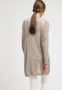 Beige long-sleeve cardigan with a textured knit pattern, featuring a loose fit and side slits, worn over white pants.