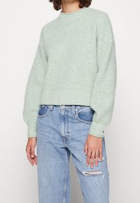 Jasnozielony, teksturowany sweter z dzianiny z okrągłym dekoltem i bufkami, połączony z jasnoniebieskimi, przetartymi jeansami. Krótkie kroje.