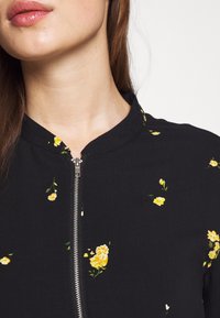 Černý květinový zipový top s žlutými květinovými vzory a stojáčkem. Má stříbrný zip a hladkou texturu látky.