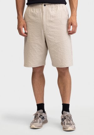 Homme portant un short beige texturé, des chaussettes noires et des chaussures de sport beige, debout devant un fond uni.