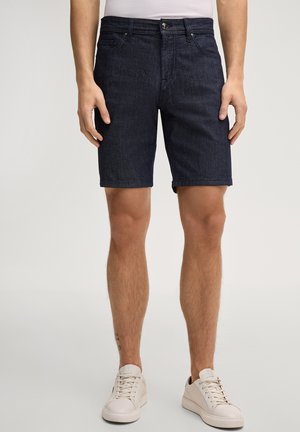 Męski model w ciemnych szortach dżinsowych i białych trampkach, stojący na tle jednolitego, jasnego tła, ręce swobodnie opuszczone wzdłuż ciała.