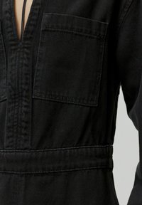 Zwarte denim jumpsuit met een diepe V-hals, twee borstzakken en een strakke taille. De stof heeft een subtiele textuur en zichtbare stiksels.