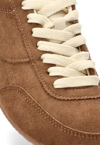Zapatilla de ante marrón con cordones de crema, presenta una punta redondeada y costuras en contraste a lo largo del costado. Textura suave, diseño casual.