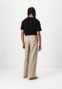 Personne aux cheveux tressés portant un t-shirt noir, un pantalon beige à jambes larges et des chaussures marron, debout face à un mur blanc.