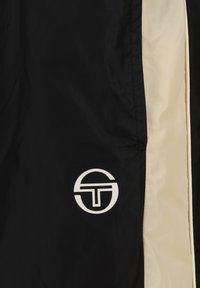 Giacca a vento nera con un pannello laterale color crema e un logo bianco prominente. Tessuto morbido e leggero.