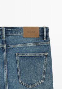 Retro dei jeans in denim blu con una tasca singola cucita e una patch in pelle marrone etichettata "Massimo Dutti" sulla vita.