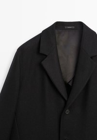 Cappotto nero in tessuto strutturato, con revers a lancia, chiusura a un bottone e foderato liscio. Nessun motivo o dettaglio visibile.