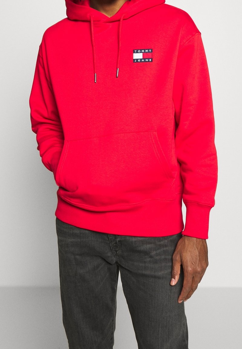 Felpa rossa con cappuccio, tasca a marsupio frontale, cappuccio con lacci e una toppa rettangolare con la scritta "TOMMY JEANS". Tessuto misto cotone.