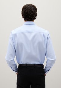 Man met donker golvend haar, in een lichtblauw geruit overhemd dat in zwarte nette broek is gestoken, met de rug naar een plain muur gekeerd.