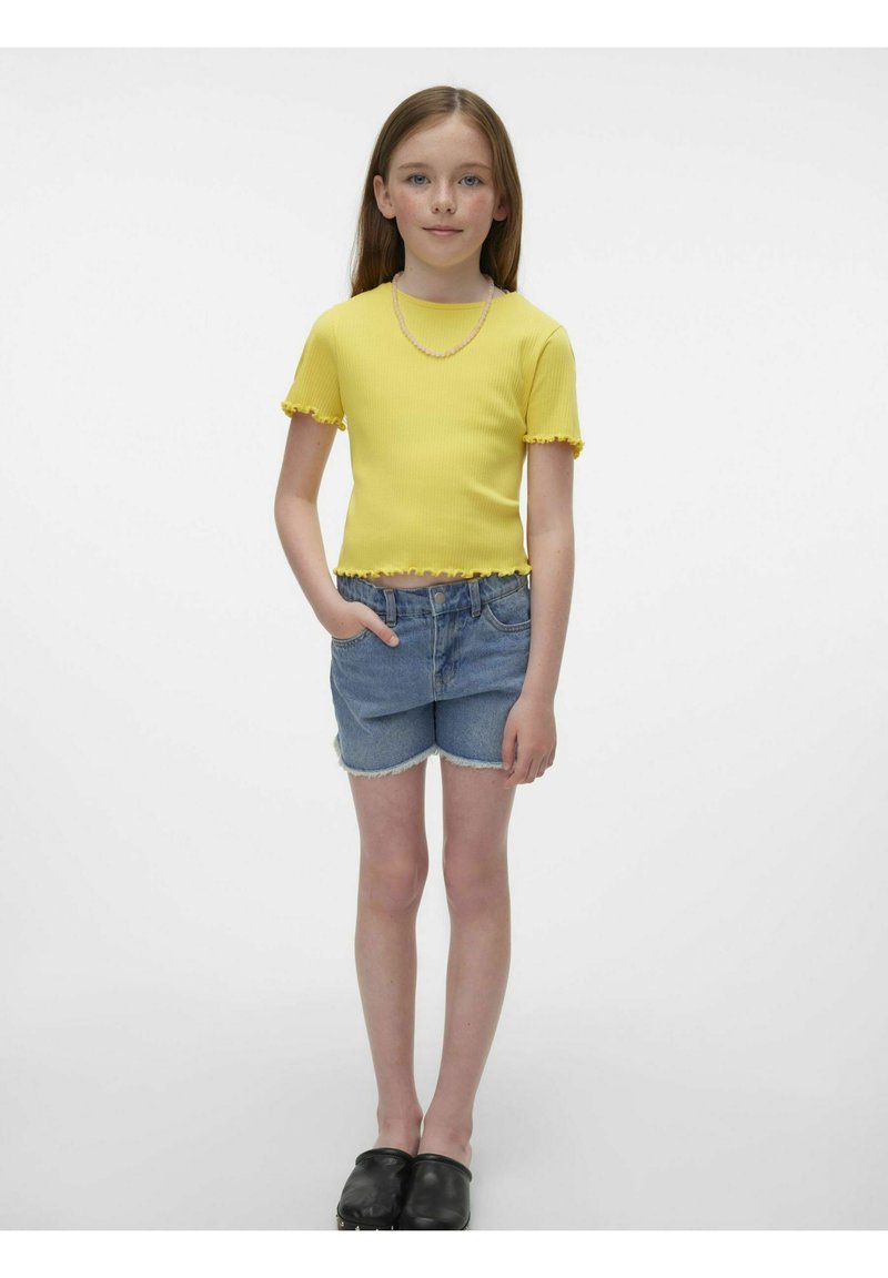 Top corto giallo a coste con bordi arricciti, abbinato a pantaloni shorts in denim blu chiaro con orli sfilacciati. Scarpe slip-on nere.