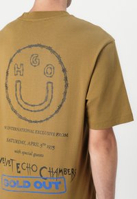 Olive green cotton t-shirt featuring a barbed wire design with a smiley face, black text, and a blue "SOLD OUT" label on the back.