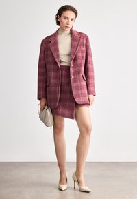 Red and pink plaid blazer with matching mini skirt, beige turtleneck, and cream high-heeled shoes, holding a beige clutch bag.